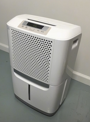 A white dehumidifier sitting on a gray floor up against a white wall. 