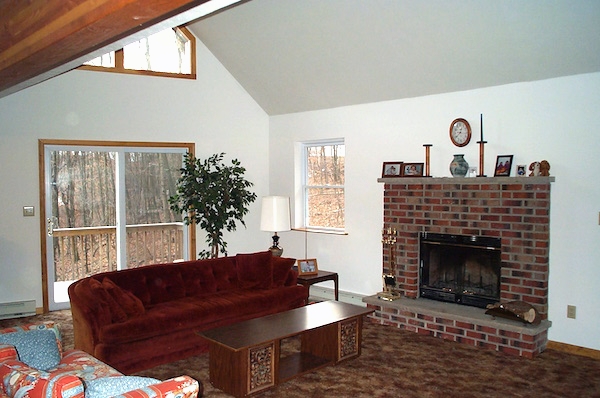 A living room with a fireplace, rust colored couch, red and blue love seat, wooden coffee table, tan carpets and large windows letting a lot of light in.