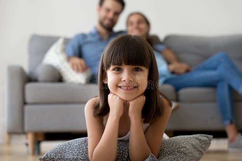 Adorable little girl looking at camera lying on warm floor. Relaxing with parents at home, smiling happy child posing playing in living room on family weekend stock photo