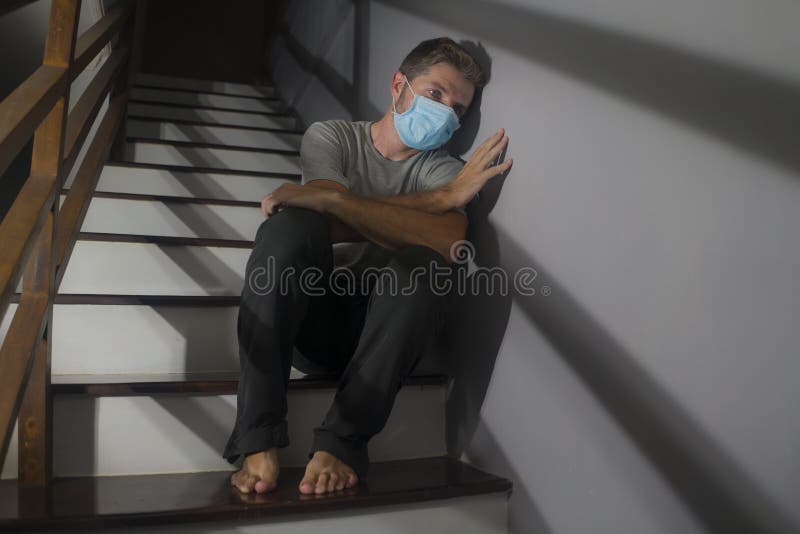 Dark portrait of young scared and worried man in protective mask sitting on stairs at home staircase during lockdown and. Quarantine for covid-19 virus pandemic royalty free stock photography