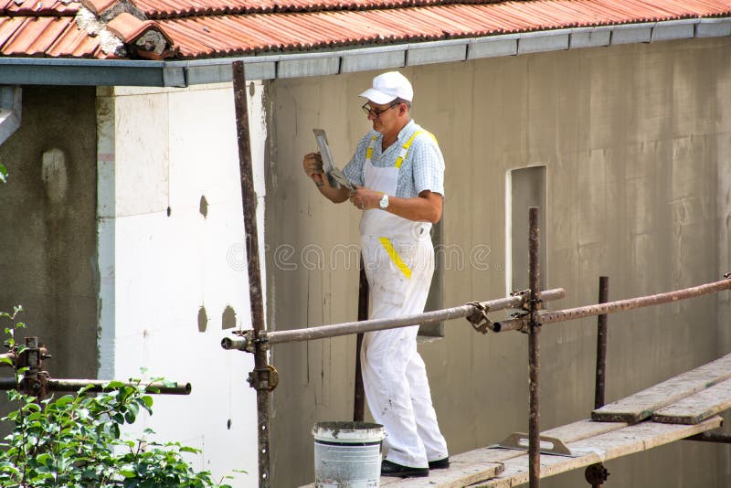 Facade Plasterer with white uniform is on scaffold. Nis, Serbia - June 16, 2018: Facade Plasterer man with white uniform is on scaffold. Worker renovates stock photos