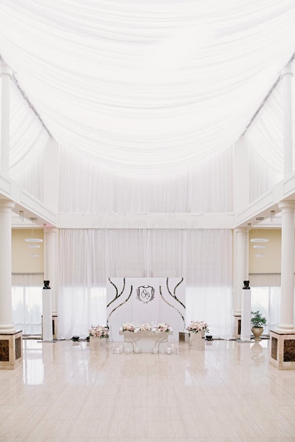 Impressive large wedding hall with high white ceilings, bright parquet and decorated table for groom and bride. Luxurious decor in white tones with silver stock images