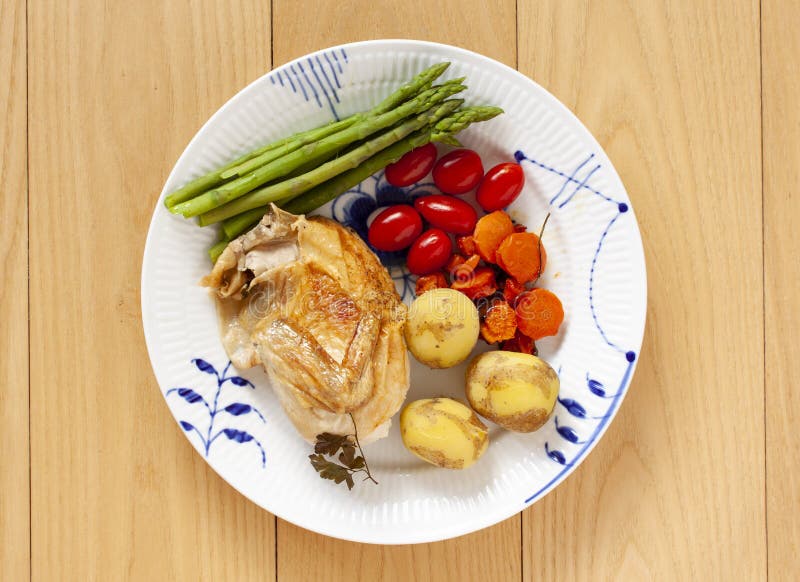 Oven-roasted chicken with plum tomatoes, new potatoes and green asparagus on a plate. Top view on wooden background. - Image royalty free stock photo