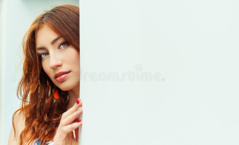 beautiful girl with red hair and full lips peeping from behind the white wall stock image
