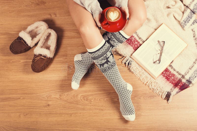 Young woman sitting on the wooden floor with cup of coffee, plaid, cookie and book. Close-up of female legs in warm socks with a. Soft photo of woman on the bed stock photo