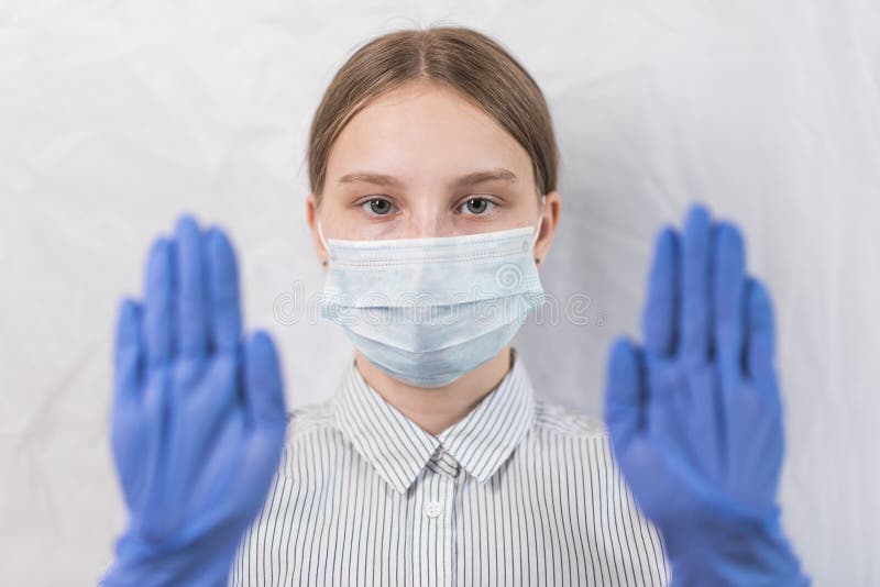 Teen girl 12-15 years old, in medical mask covers her face, hand gesture with gloves on feet, attention dangerous, white. Background, COVID-19 sars-cov-2 stock image