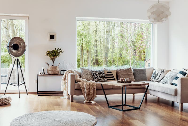 View outside to the green woods through large glass windows in a natural living room interior with beige sofa and dark hardwood fl. Oor concept stock photos