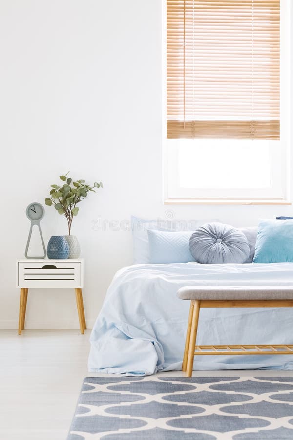 Window with wooden blinds in white bedroom interior with bed wit. H light blue bedclothes, carpet with pattern and bedside table with clock and plant royalty free stock photography