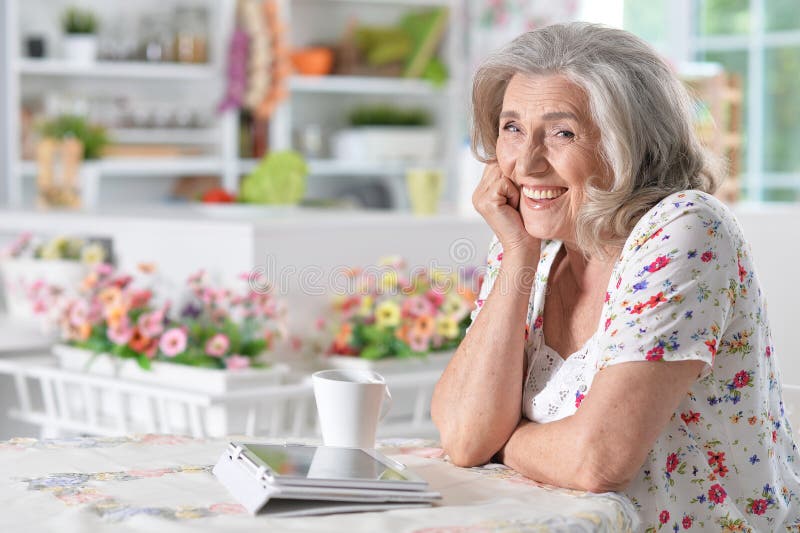 Woman using digital tablet. Beautiful senior woman smiling at camera while drinking tea at table with digital tablet on it stock image
