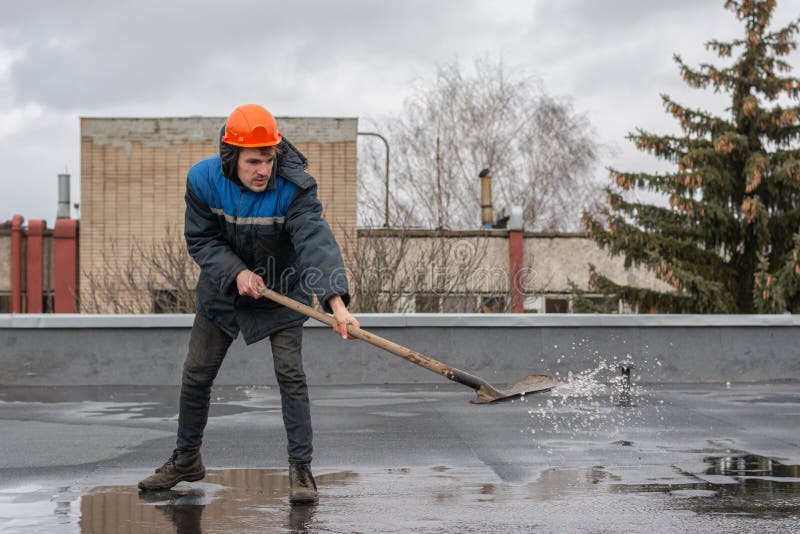 Worker builder on the roof of the factory removes water from the puddle with a shovel. on the head a construction helmet. stock photography