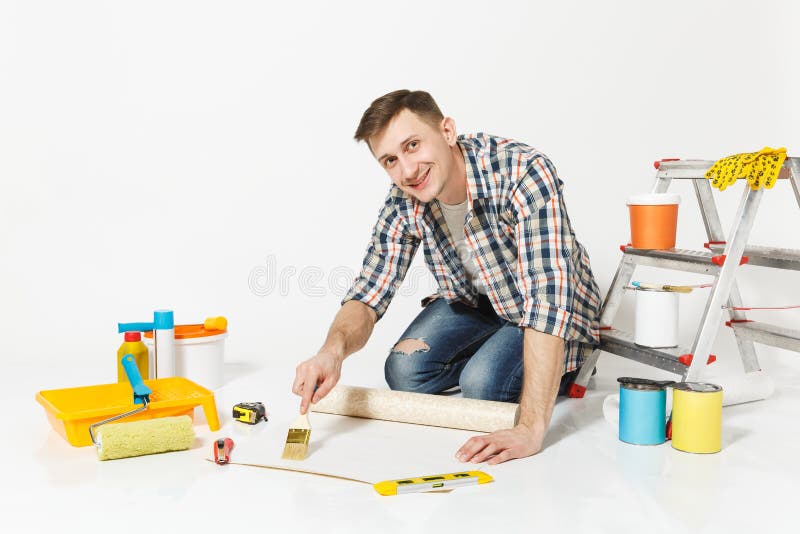 Young man sitting on floor with wallpaper roll, brush for gluing, instruments for renovation apartment room isolated on stock photography