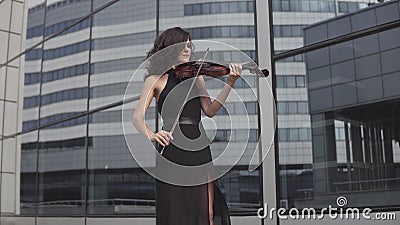 Elegant woman in black dress playing violin near glass building. Art concept stock video footage