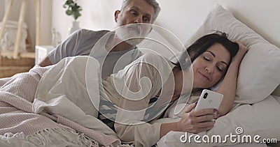 Jealous senior Caucasian man looking over shoulder of wife at smartphone screen as woman messaging with someone stock video